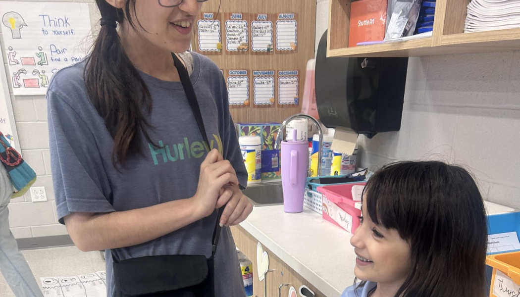 A smiling teacher talks warmly with a young student in a brightly lit elementary classroom.