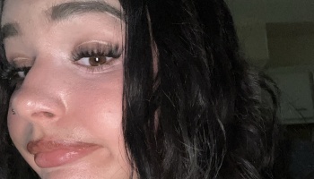 Close-up selfie of a young woman with dark wavy hair, long eyelashes, brown eyes, and a nose piercing.