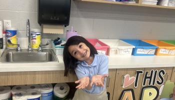 A smiling young student in a school uniform holds out her hands in front of classroom storage bins and a sink.
