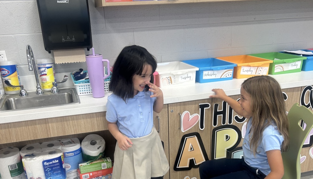Two smiling elementary school girls in light blue uniforms laughing together in a classroom.