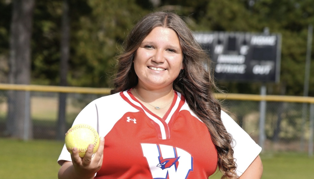 A smiling young softball player in a red and white jersey holding a yellow softball on a field.