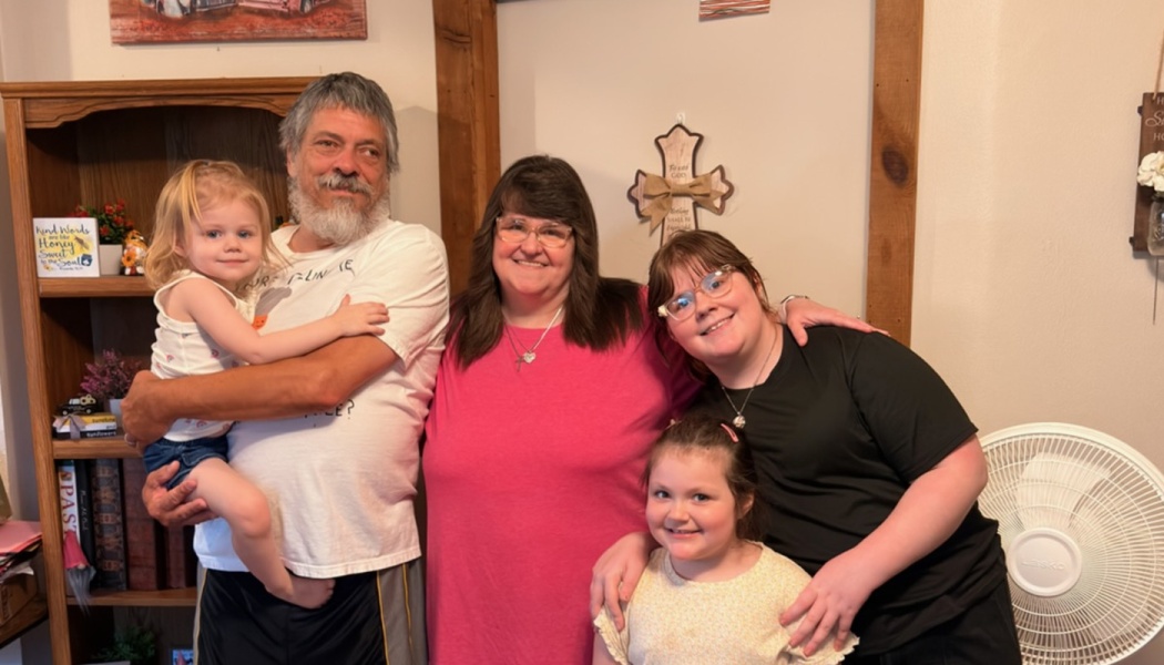 A smiling family of five standing closely together indoors, with the father holding a young toddler.