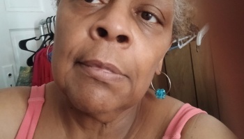 Close-up portrait of a woman with grey hair, wearing a pink tank top and a blue hoop earring.
