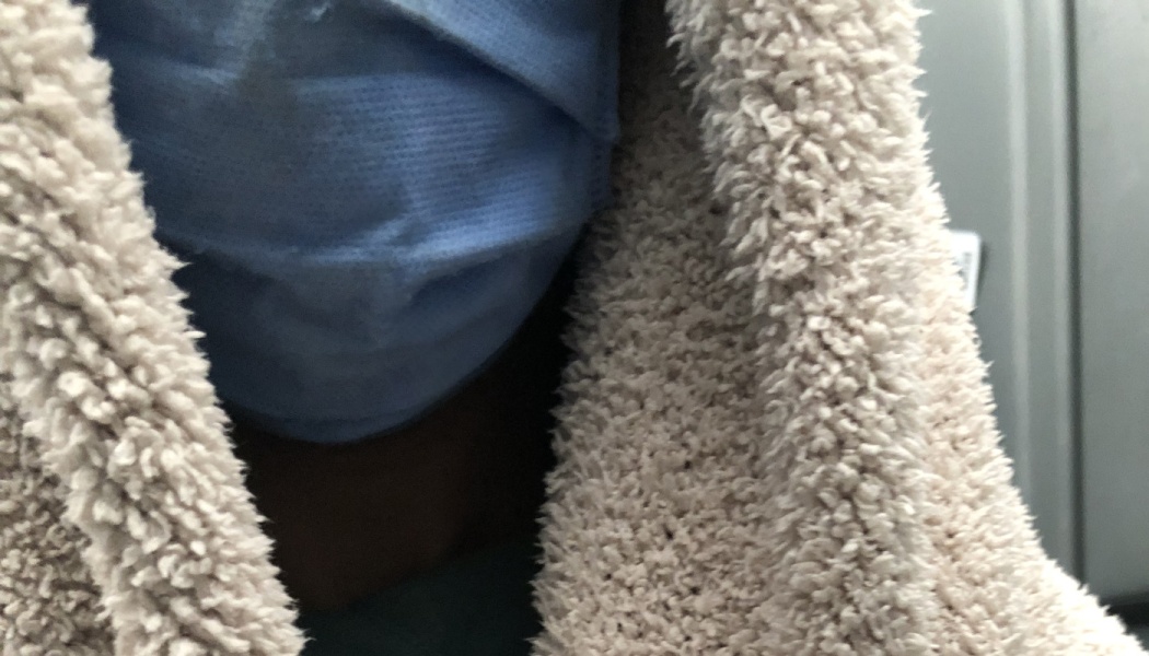 Close-up of a person wearing a blue surgical mask and wrapped in a fuzzy beige blanket.