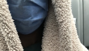 Close-up of a person wearing a blue surgical mask and wrapped in a fuzzy beige blanket.