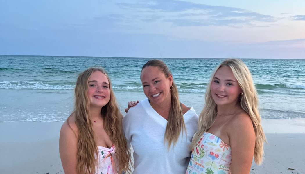 A mother and her two daughters smile together on a sandy beach with ocean waves in the background.