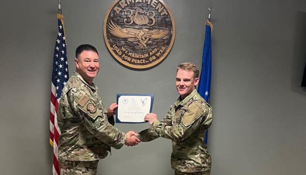Two military members in camouflage uniforms shake hands while holding a certificate between two flags.