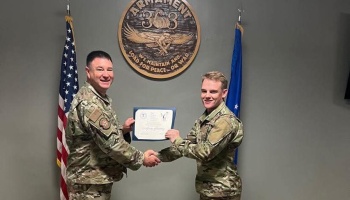 Two military members in camouflage uniforms shake hands while holding a certificate between two flags.
