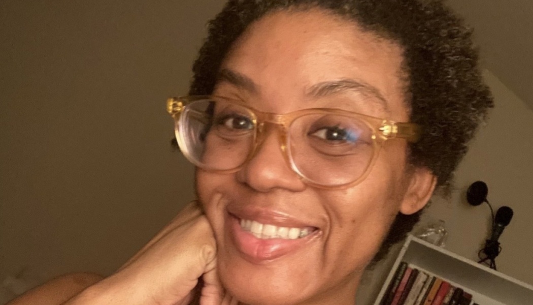 A smiling Black woman with short natural hair and clear glasses resting her chin on her hand.