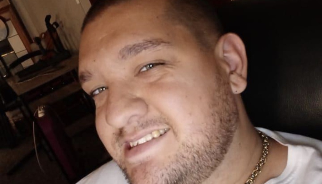 Close-up portrait of a smiling young man with short hair, facial stubble, light eyes, and a gold chain necklace.
