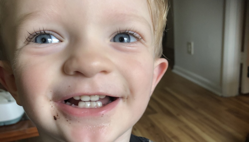 Close-up of a smiling blonde toddler with bright blue eyes and chocolate smudges around their mouth.