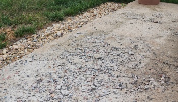Severely cracked and crumbling concrete walkway in need of repair, bordered by small rocks and grass.