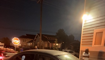 A residential neighborhood street at night with an illuminated stop sign, parked cars, and dimly lit houses.