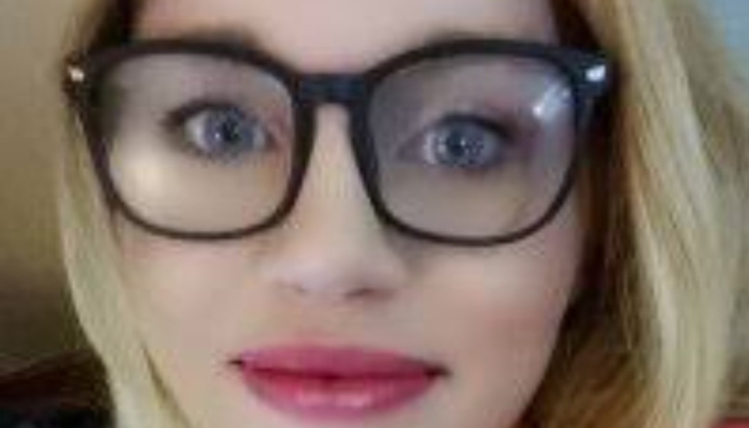 Close-up of a blonde woman wearing black-framed glasses and pink lipstick looking at the camera.