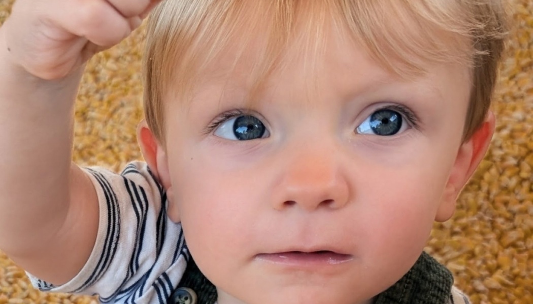 Close-up of a blonde toddler with wide blue eyes looking up while reaching a hand toward the camera.