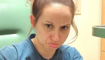 A young woman with hair in a messy bun looking tired and sad while sitting in a green medical chair.