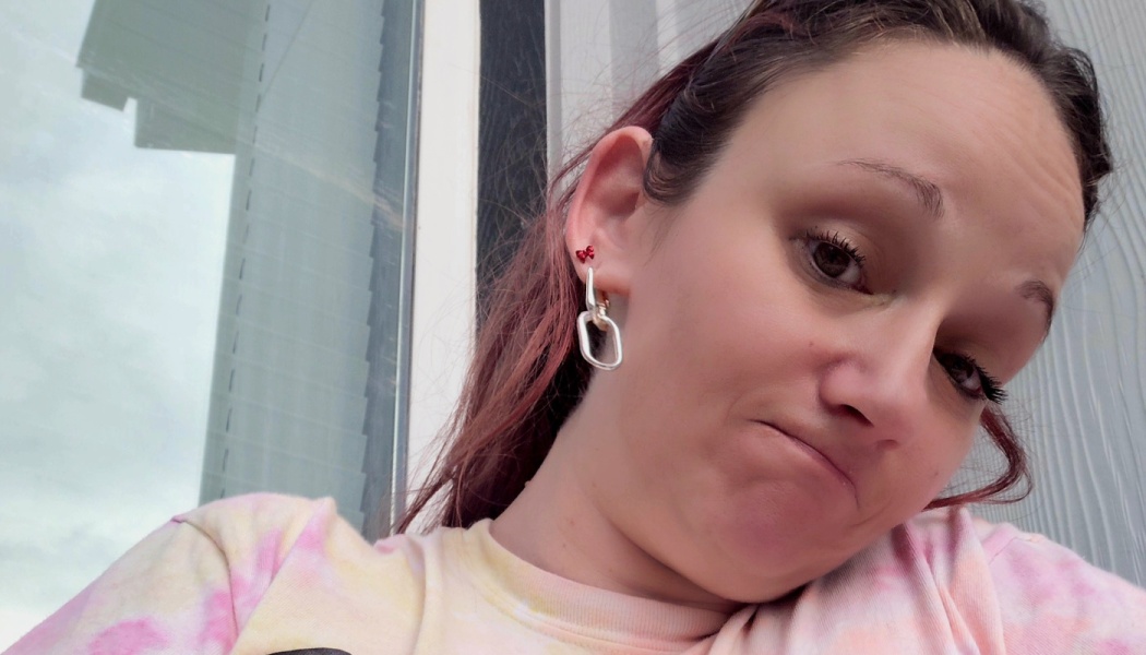 Woman with reddish hair and silver chain earrings wearing a pastel tie-dye shirt looking at the camera.