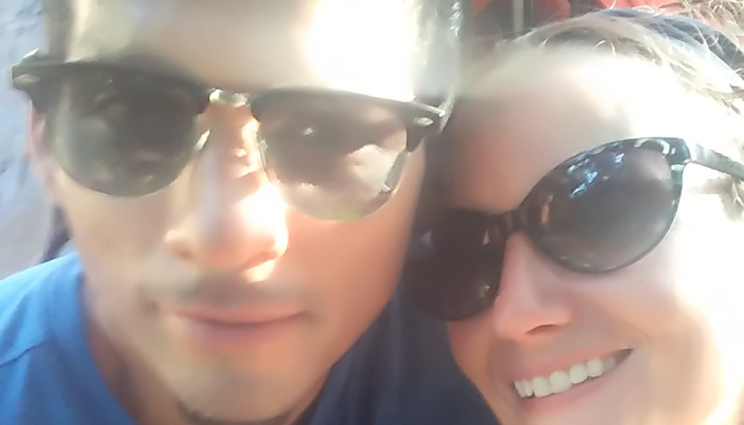 Close-up selfie of a man and a smiling woman, both wearing dark sunglasses in bright sunlight.