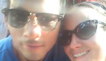 Close-up selfie of a man and a smiling woman, both wearing dark sunglasses in bright sunlight.