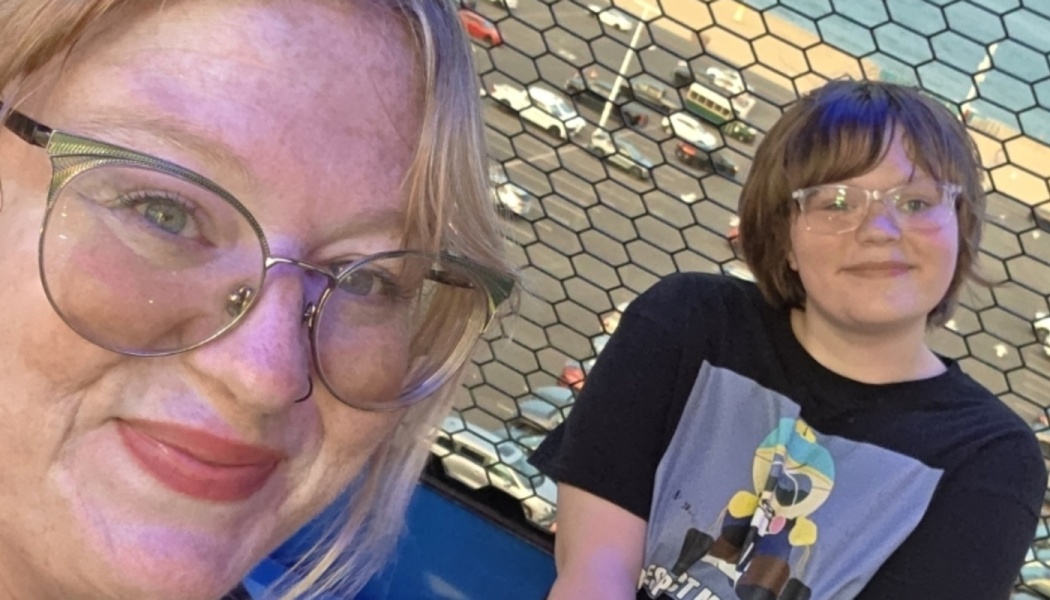 Selfie of a smiling blonde woman with glasses and a young person on a ride overlooking a city street.
