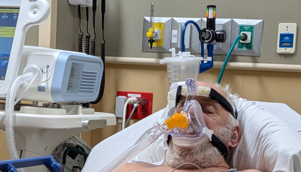 An elderly man with a white beard lies in a hospital bed wearing a clear oxygen mask attached to a ventilator.