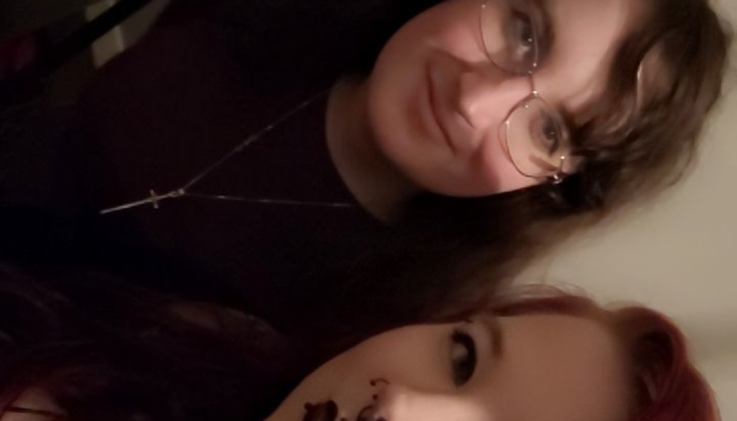 Two young women posing close together, one wearing round glasses and a cross necklace, alongside a red-haired woman.