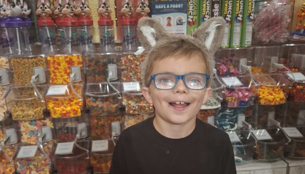 A smiling boy wearing glasses and plush wolf ears stands in front of colorful candy store bins.
