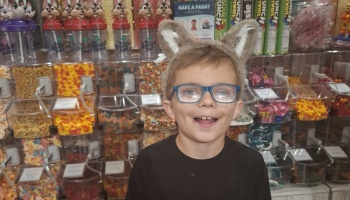 A smiling boy wearing glasses and plush wolf ears stands in front of colorful candy store bins.