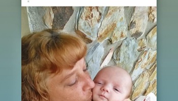 A woman with reddish-blonde hair tenderly kissing a baby's cheek against a stone wall background.
