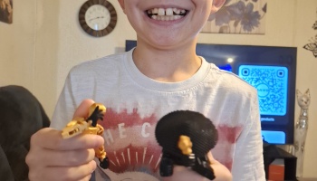 A smiling child holds up two small action figures, one gold and one black.