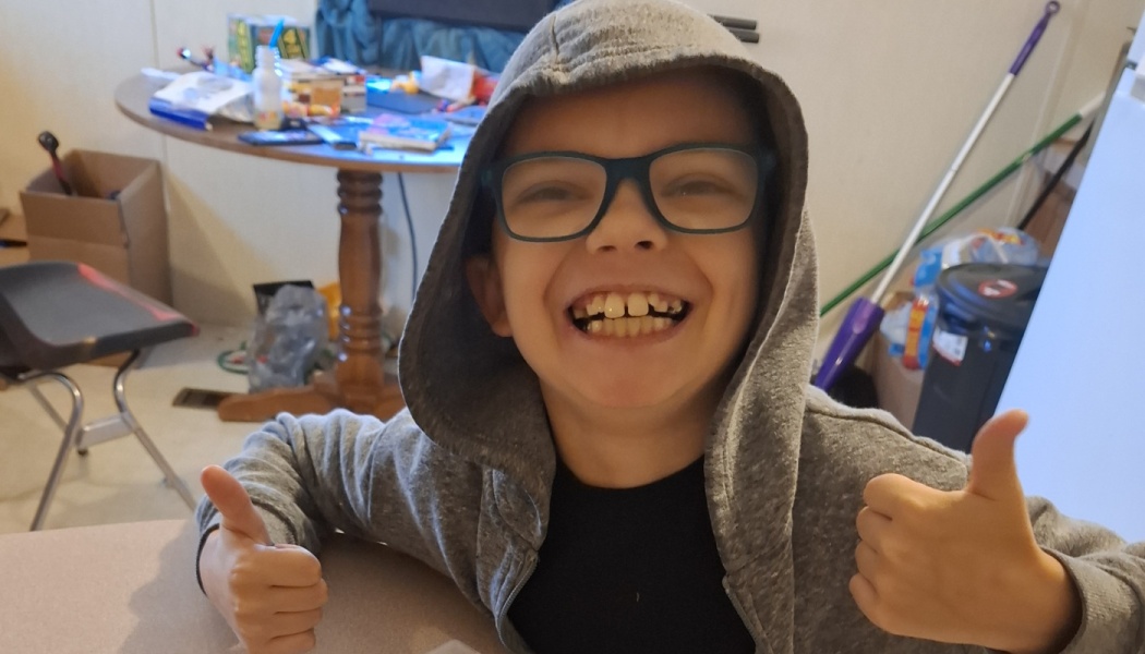 A young boy with glasses wearing a grey hoodie smiles widely while giving two thumbs up.