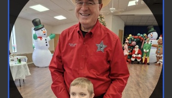 A smiling sheriff in a red shirt stands behind a young boy at a festive Christmas event.