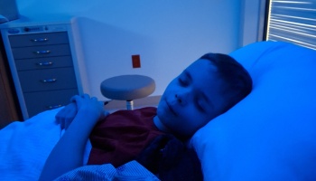 Young boy sleeping peacefully in a hospital bed illuminated by dim blue lighting.