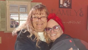 Two smiling women hugging, one with long blonde hair and glasses, the other wearing glasses and a red headwrap.