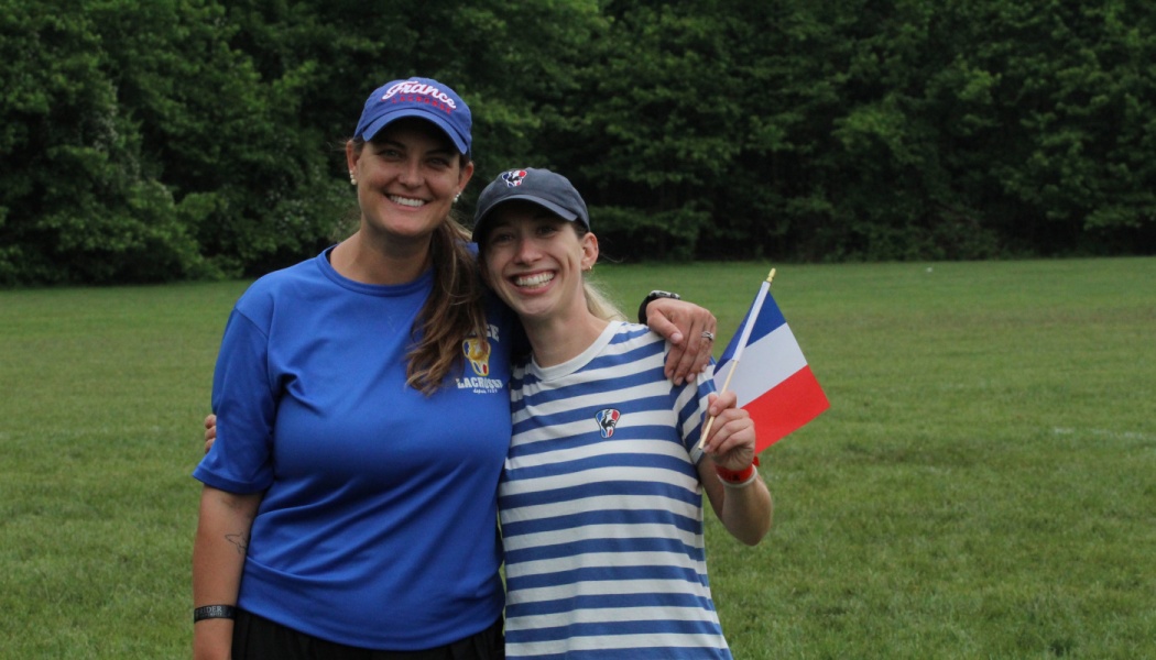 Two smiling women wearing France Lacrosse apparel pose on a grassy field, one holding a small French flag.