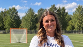 A smiling woman in a white France shirt stands on a lacrosse field with an orange net behind her.