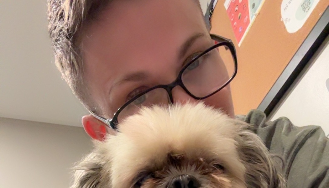 A person wearing black-rimmed glasses poses closely behind a small, fluffy dog.