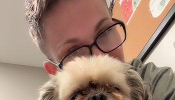 A person wearing black-rimmed glasses poses closely behind a small, fluffy dog.