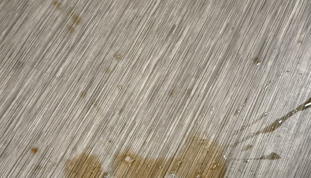 Spilled brown liquid and droplets pooling on a light grey textured wood-grain surface.