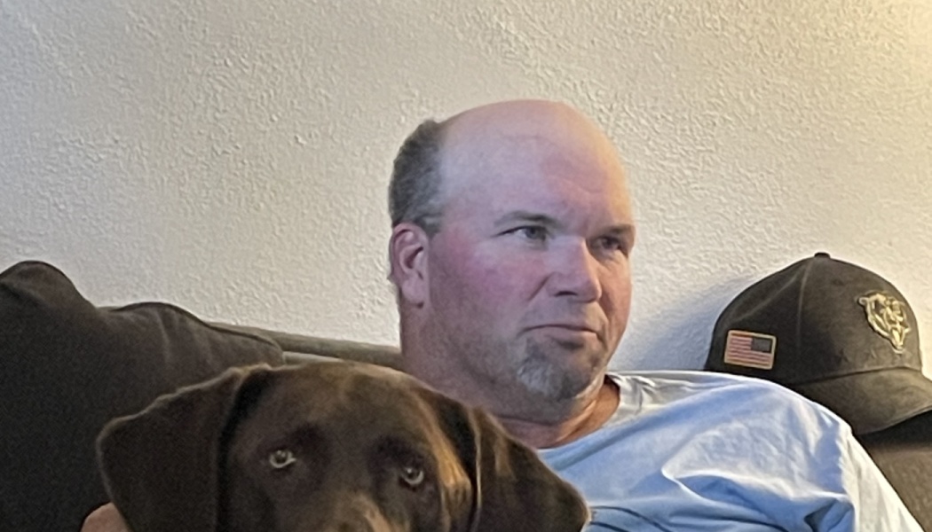 A man in a blue t-shirt sitting on a couch next to his chocolate Labrador.