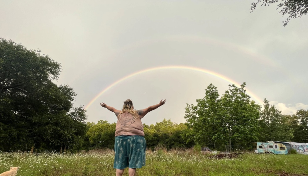 A person stands in a grassy field with arms outstretched toward a vibrant double rainbow in the sky.