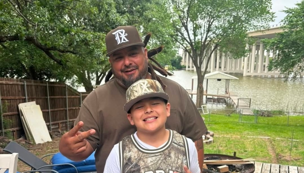 A smiling man throwing a peace sign stands behind a young boy wearing a camo cap outdoors near water.