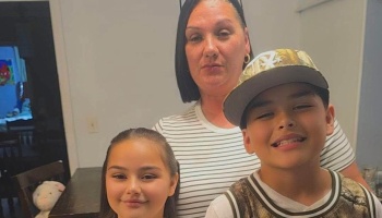 A mother in a striped shirt poses indoors with her young daughter and smiling son wearing a camo baseball cap.