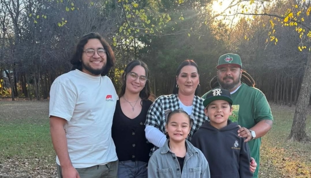 A smiling family of six poses together outdoors in a park with trees in the background.