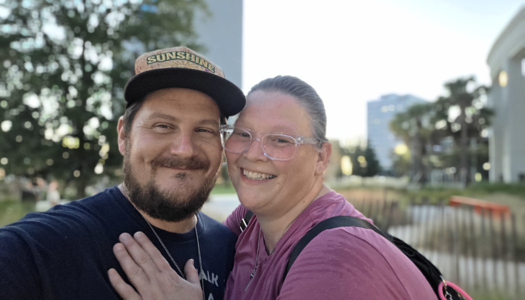 A smiling couple posing for a selfie outdoors, a bearded man wearing a hat beside a woman with clear glasses.