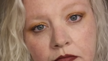 Close-up of a woman with curly blonde hair, gold glitter eyeshadow, and dark red lipstick.