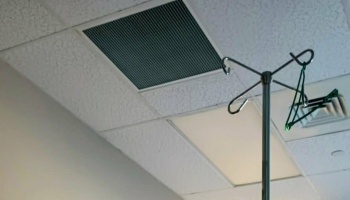 An empty metal IV pole in a hospital room, viewed from below against a tiled ceiling with air vents.
