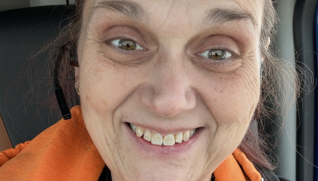 Close-up selfie of a smiling woman wearing an orange hoodie and a phone headset.