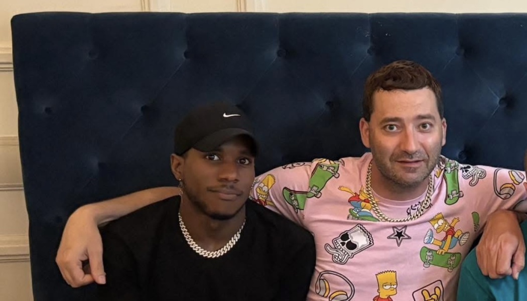 Two men sitting on a dark blue tufted couch, one in a black Nike cap and the other wearing a pink Bart Simpson shirt.