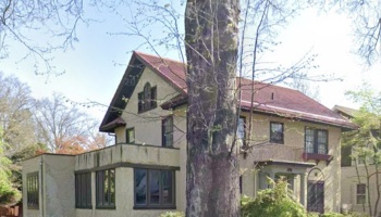 Historic two-story stucco house with a red tile roof, dark-trimmed windows, and a large tree in the foreground.